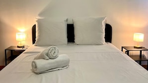 Ein ordentlich bezogenes Bett mit weißen Leinentüchern, zwei Kissen und einem zusammengerollten Handtuch. An beiden Seiten des Bettes befinden sich Nachttische mit Lampen.