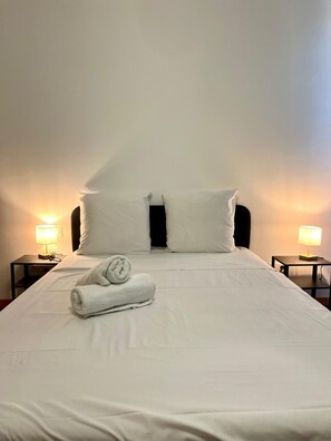 Ein ordentlich bezogenes Bett mit weißen Leinentüchern, zwei Kissen und einem zusammengerollten Handtuch. An beiden Seiten des Bettes befinden sich Nachttische mit Lampen.