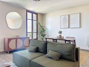 A living room with a sofa, a cabinet, a dining table, and two wall-mounted artworks. - L'Anatole - superb harbor view, hypercentre & cosy (Toulon)