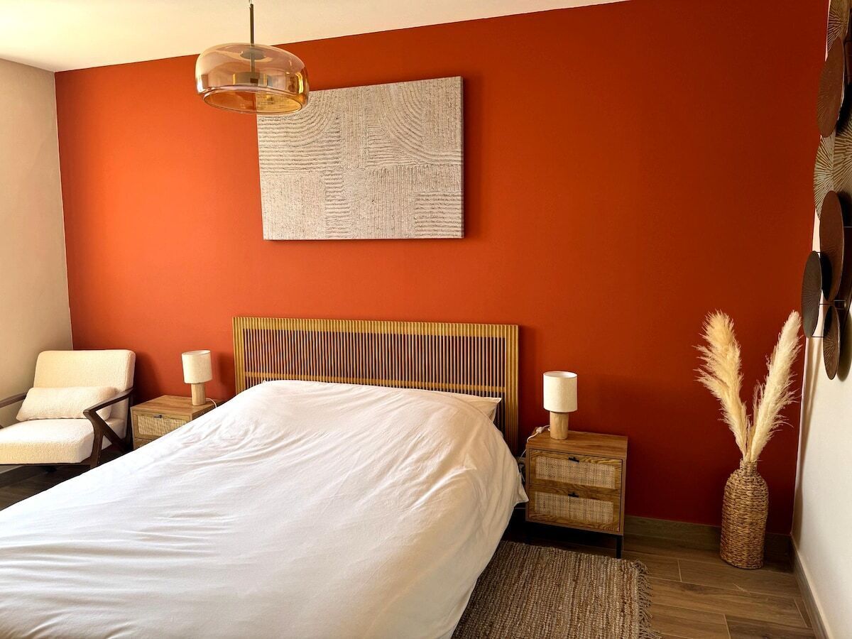 Un dormitorio con una cama, una silla, mesitas de noche, una lámpara y una obra de arte en la pared.