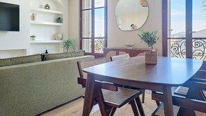 Uma sala de jantar com mesa e cadeiras de madeira, um sofá verde, uma televisão na parede e uma varanda com vista.