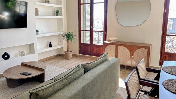 Uma sala de estar com sofá, televisão, estantes, uma planta em vaso e um espaço de jantar com cadeiras e mesa.