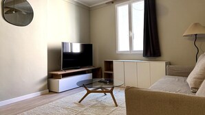 A living room with a flat-screen TV, a glass coffee table, a sofa, a lamp, a window with curtains, and a radiator. - Comfortable apartment in the heart of the city (Toulon)