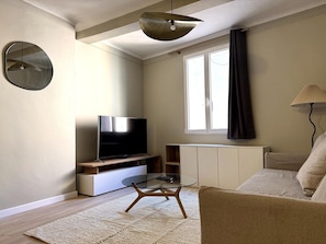 Ein Wohnzimmer mit Flachbildfernseher, Glastisch, Sofa, Lampe, einem Fenster mit Vorhängen und einer Heizung.