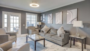 A living room with a grey sofa, a coffee table, and a lamp. There are abstract wall art pieces and a door with a view. - Poolside Fun! Waterfront Gem Near San Marco Square (Jacksonville)