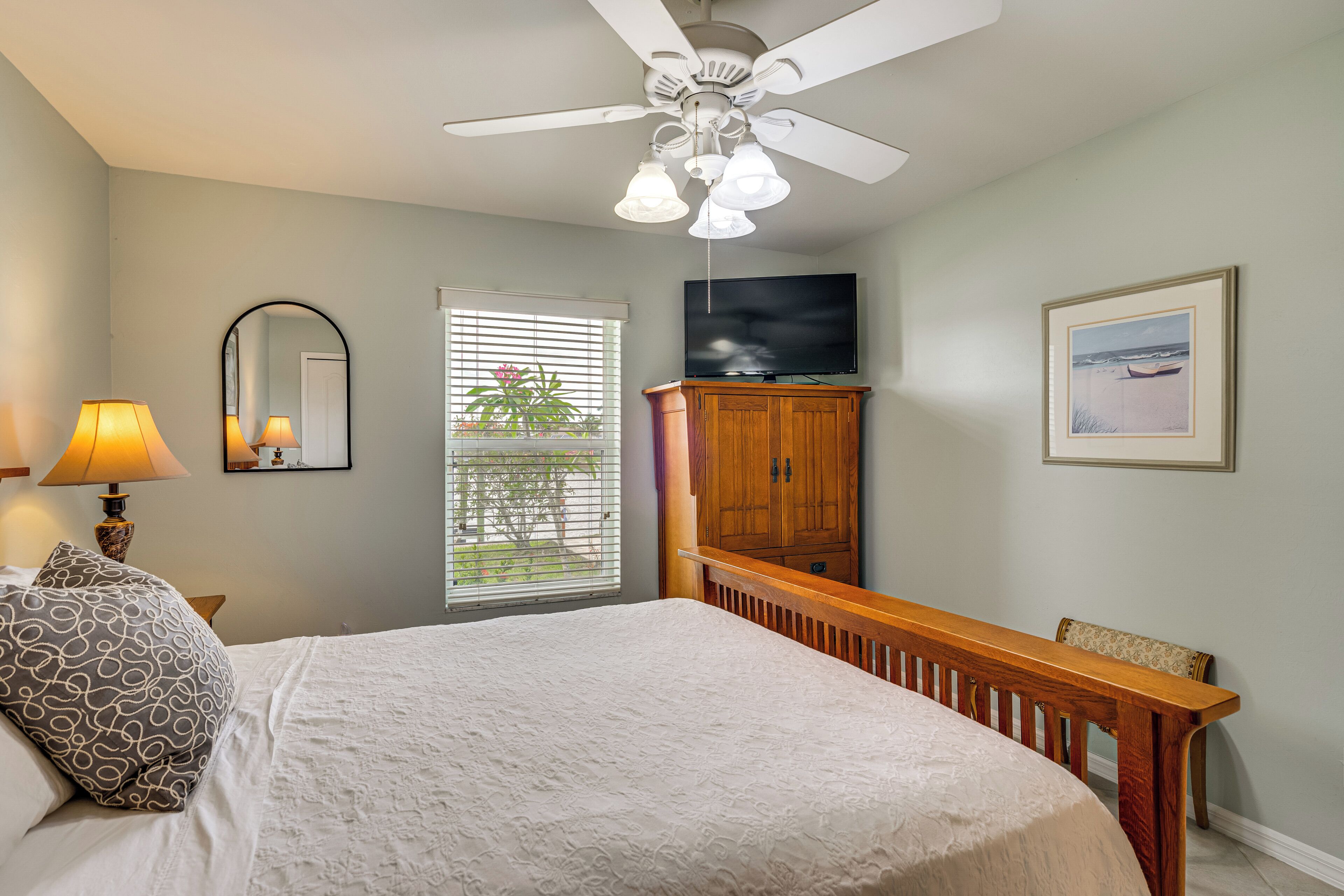 A bedroom with a bed, a ceiling fan, a TV, a wooden bench, a mirror, and a window with blinds.