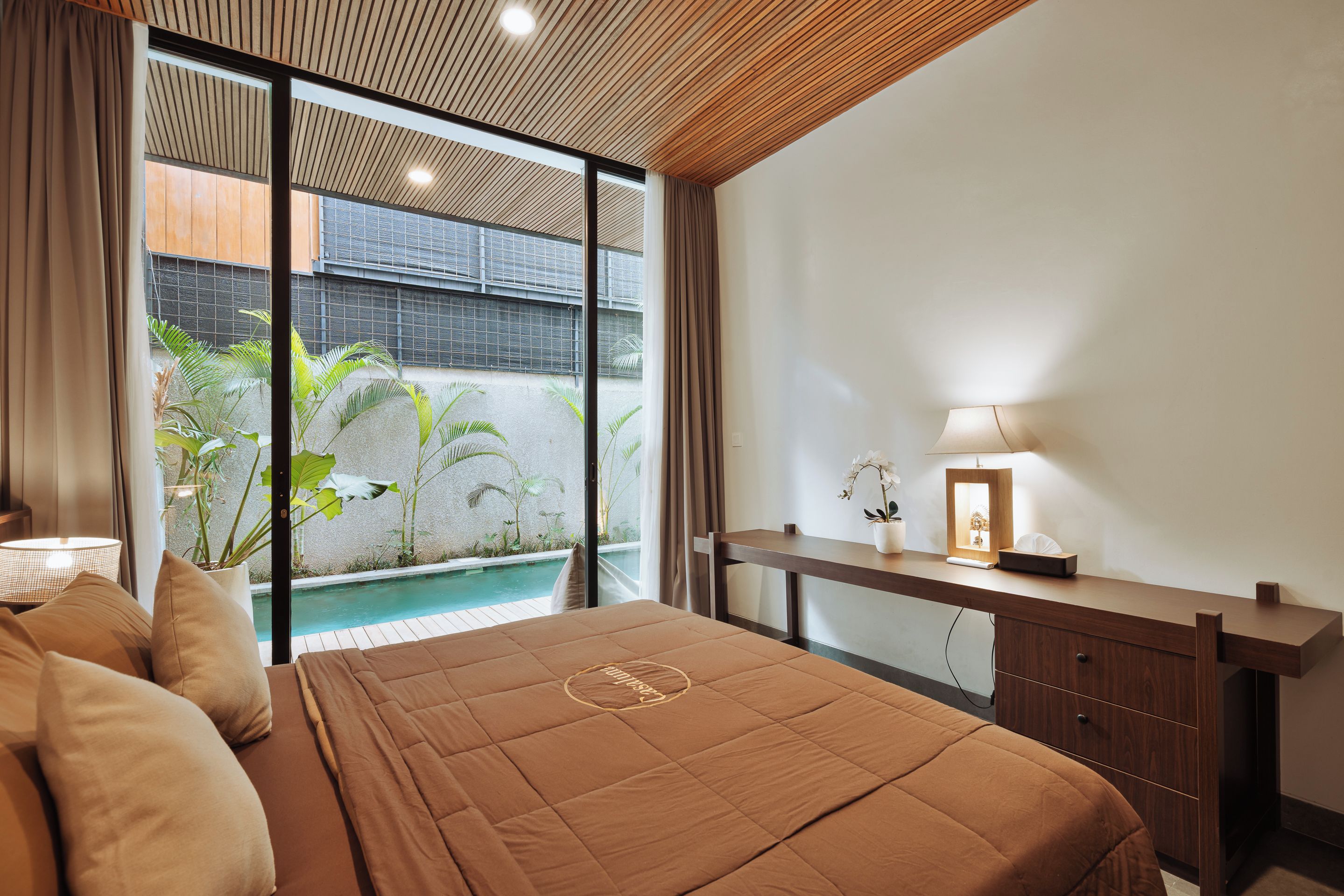 A bedroom with a bed, bedside table, lamp, and a view of an outdoor pool area.