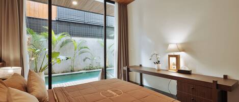 A bedroom with a bed, bedside table, lamp, and a view of an outdoor pool area.