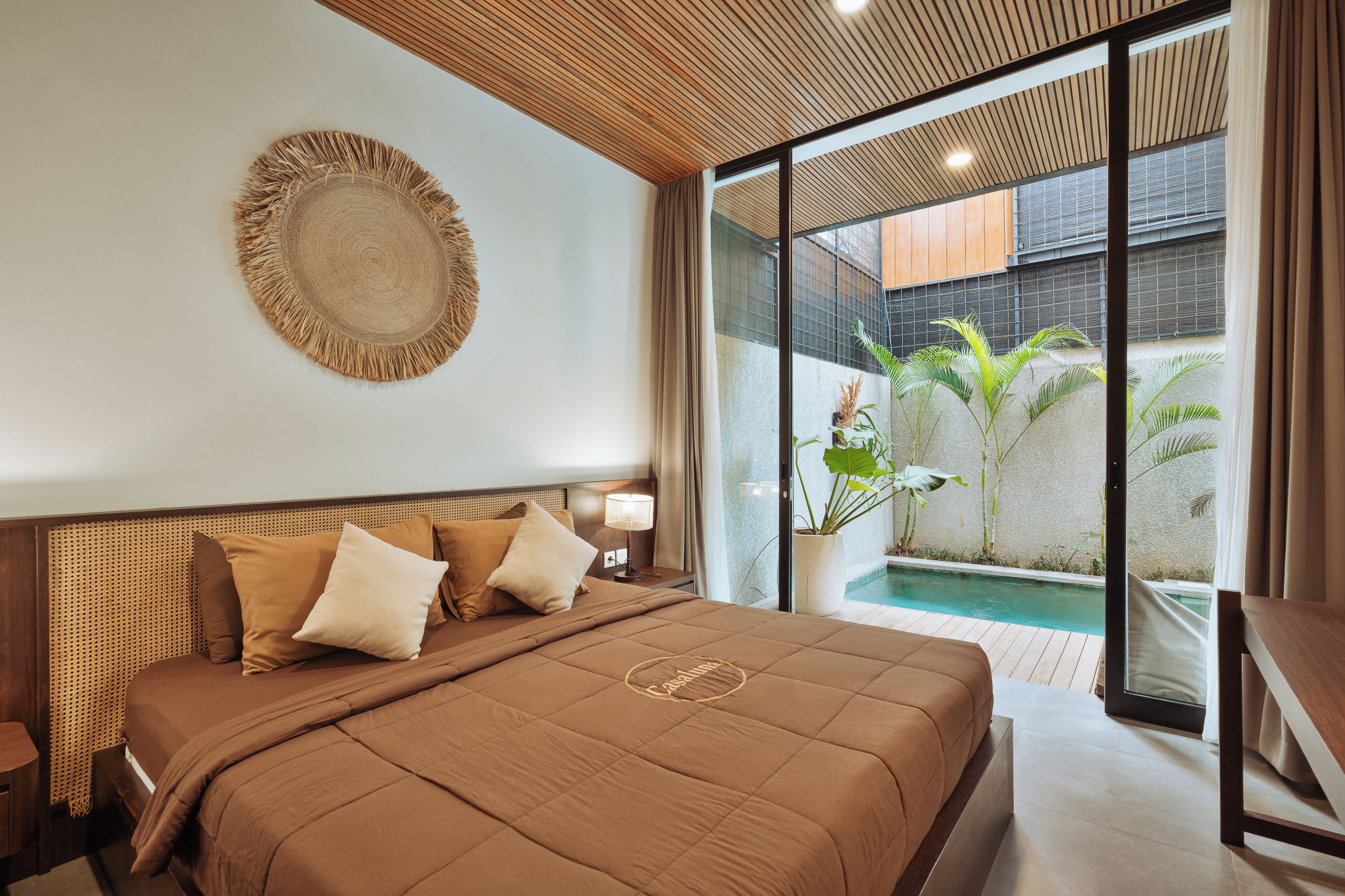 A hotel room with a large bed, a round woven wall hanging, a view of a swimming pool, and a balcony with plants.