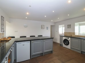 A kitchen with a built-in dishwasher, a washing machine, and a dining area with chairs. - Llanallgo Church Rooms (Moelfre)