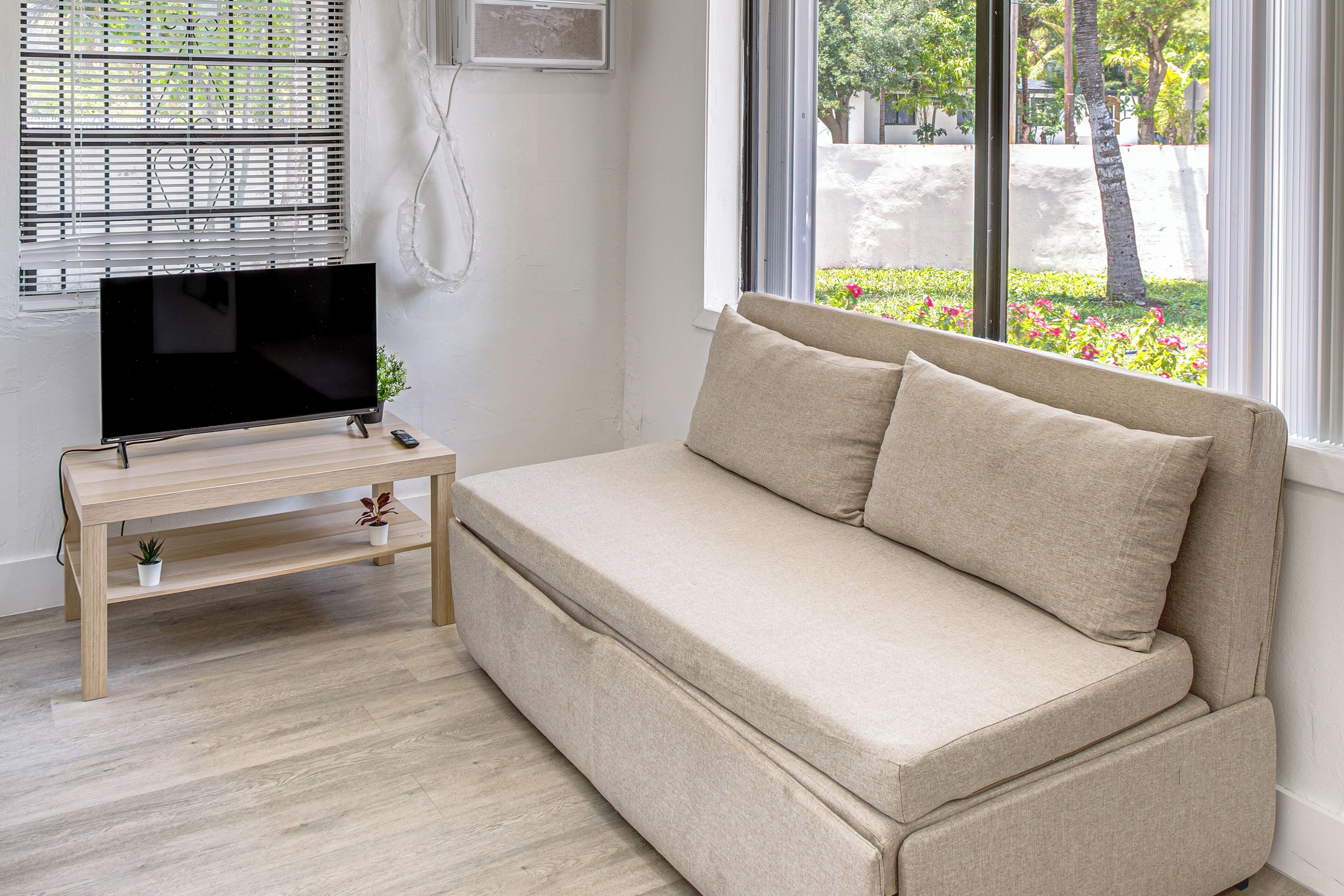 Ein Zimmer mit beiger Couch, einem Fernseher auf einem Holzboden und einem Fenster mit Blick auf Grünflächen.