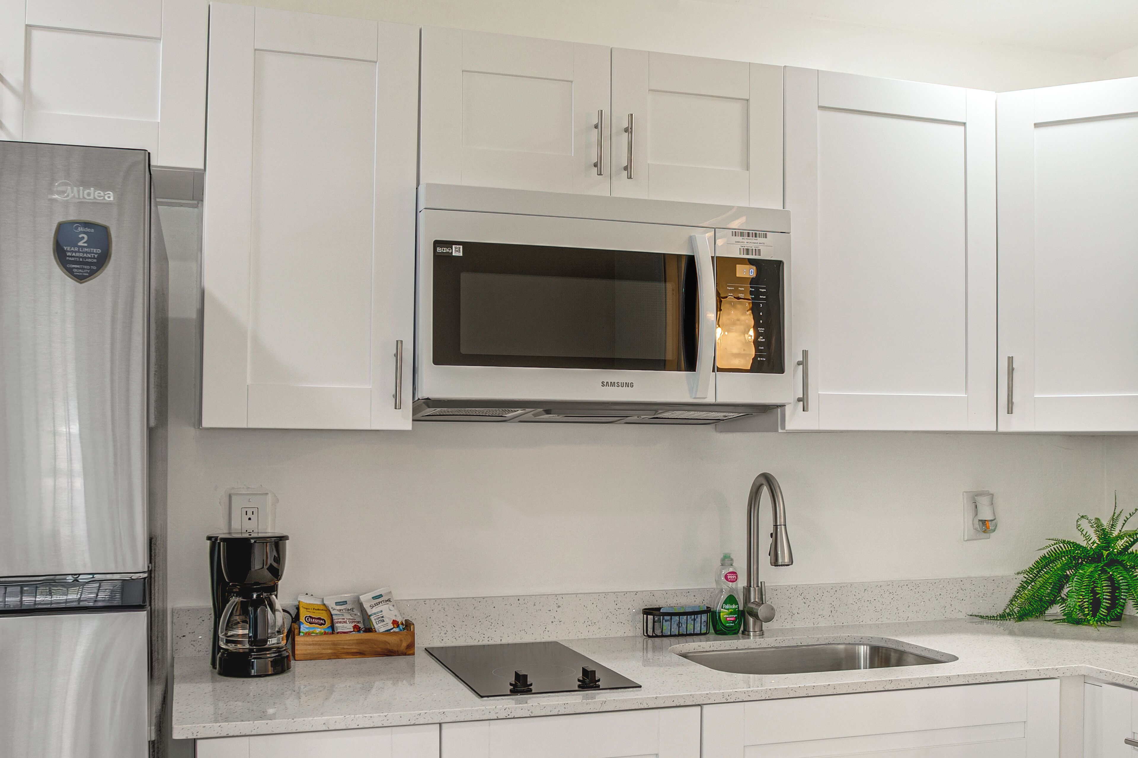 Una cocina moderna con armarios blancos, un refrigerador de acero inoxidable, un microondas Samsung, una cafetera y un lavamanos.