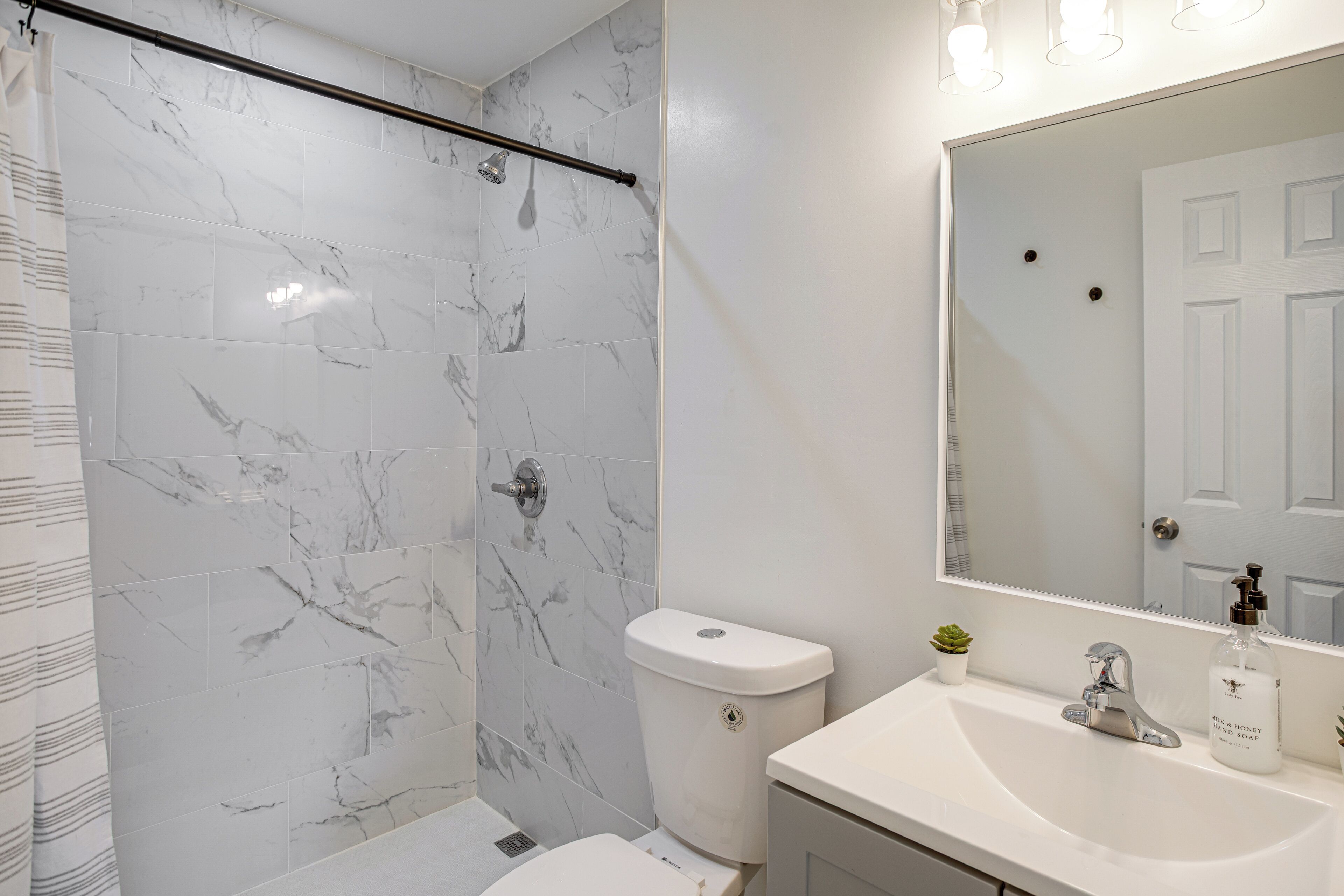 Ein Badezimmer mit einer weißen Marmordusche, einer Toilette, einem Waschbecken mit Spiegel und einer kleinen Topfpflanze.