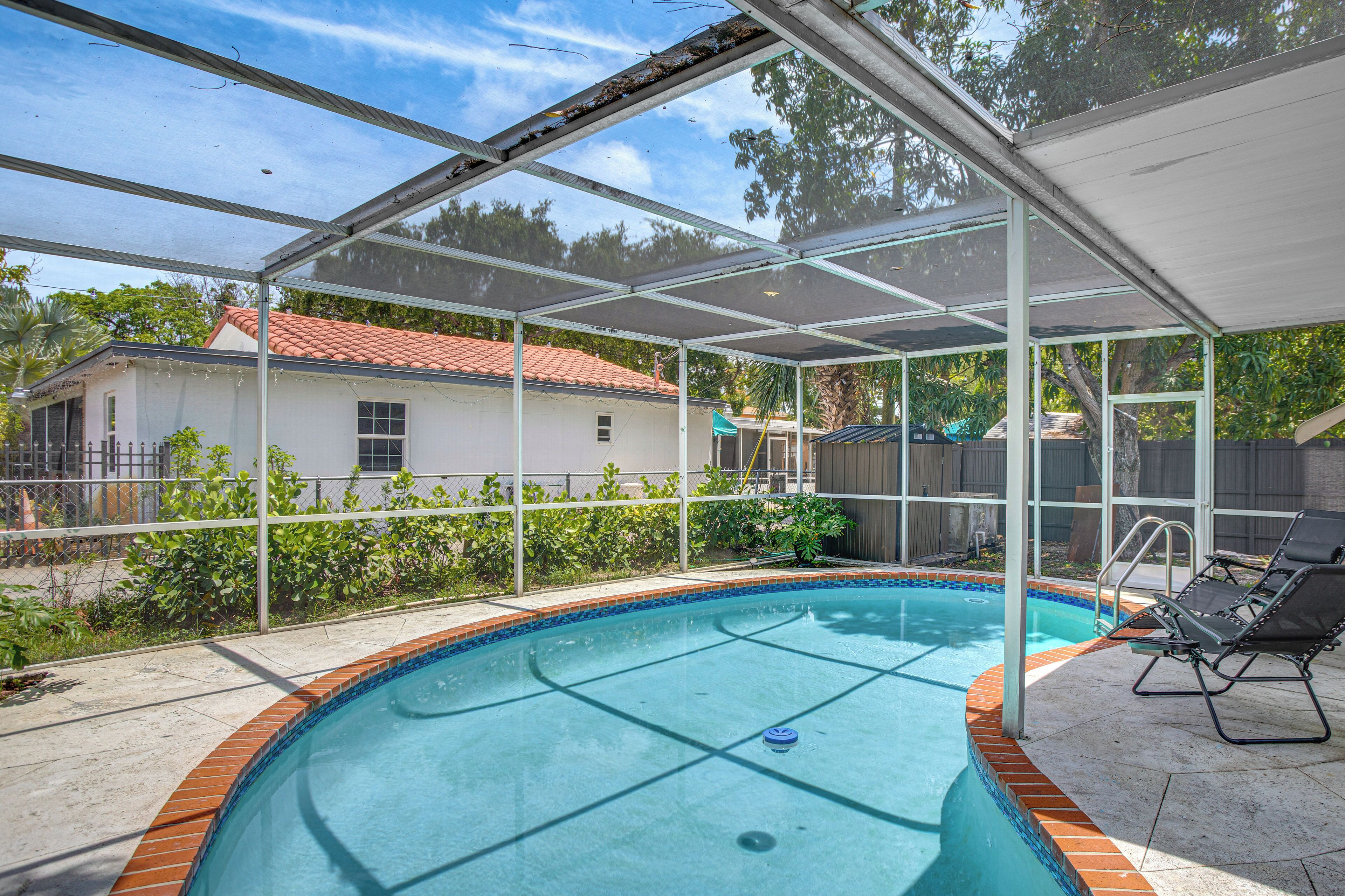 Una piscina techada con zona de patio, una casa al fondo y un jardín cercado.