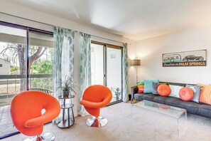 Ein modernes Wohnzimmer mit einem Glastisch, zwei orangefarbenen Sesseln, einem Sofa mit bunten Kissen und einem Schild mit der Aufschrift 'Vacation in Palm Springs'.
