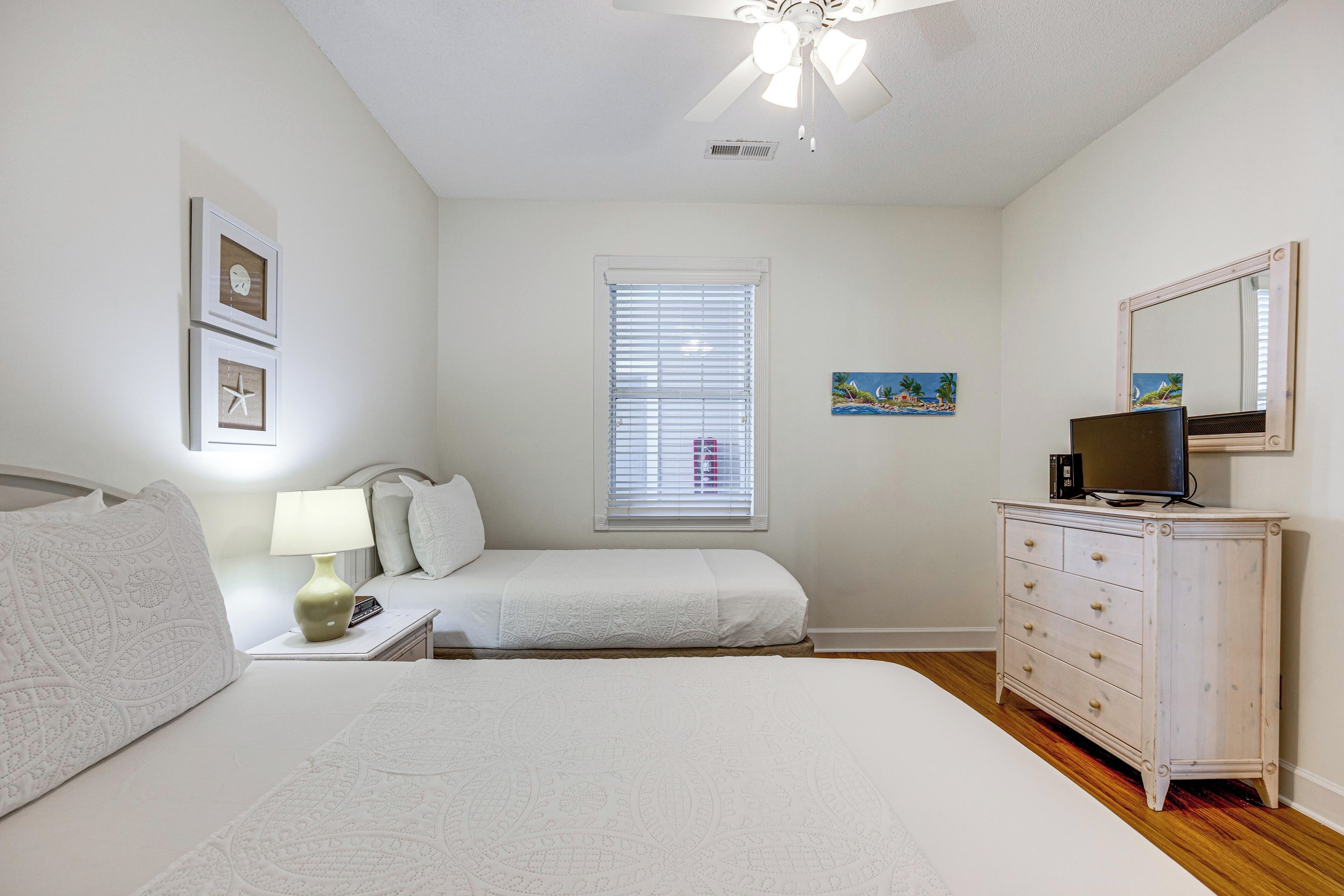 Quarto com cama, penteadeira, mesa de cabeceira, luminária, televisão, janela com persianas e ventilador de teto.