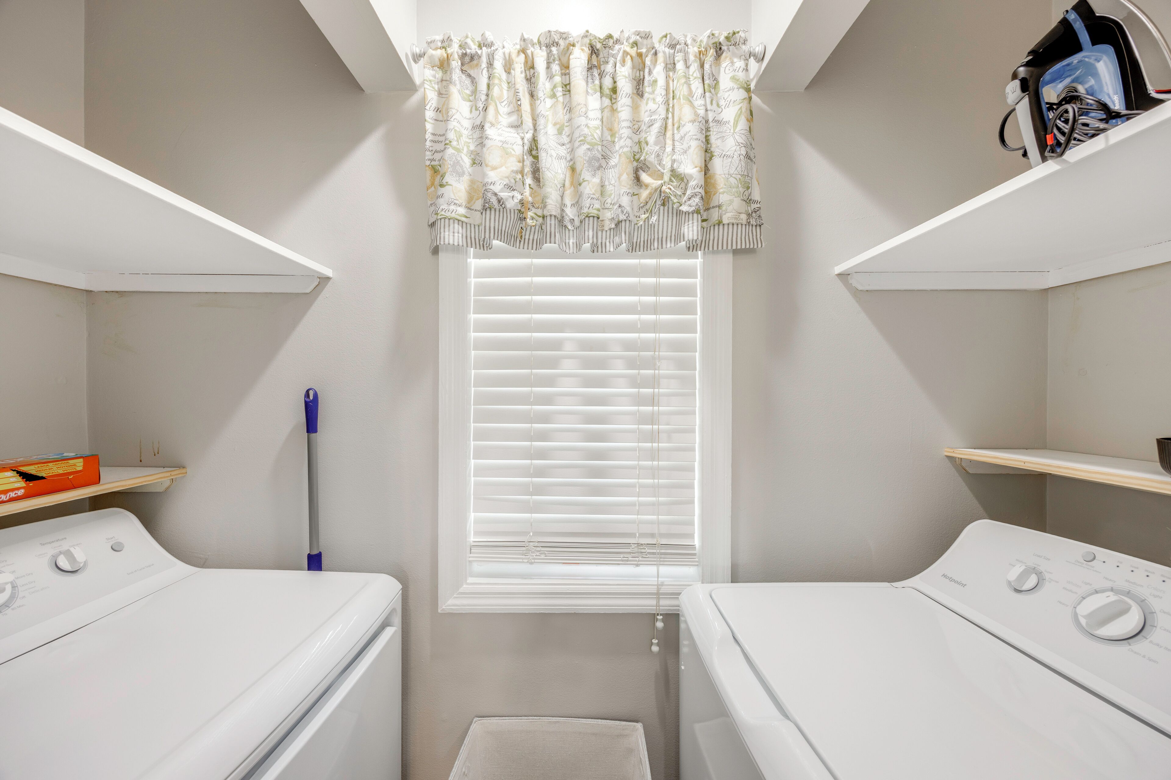A laundry room with two washing machines, a window with blinds, a mop, and a shelf with a box.