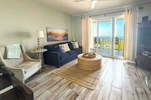 Un salón con sofá, sillón y una mesa redonda. Un cuadro adorna la pared, y hay un balcón con vistas.