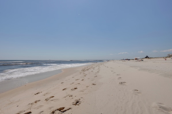 A sandy beach with footprints leading to the water, a clear sky, and distant structures.