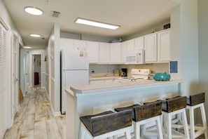 A kitchen with white cabinets, a central island with bar stools, a refrigerator, and a microwave. - Walk to Beach: The Dunes Condo w/ Resort Perks! (Gulf Shores)