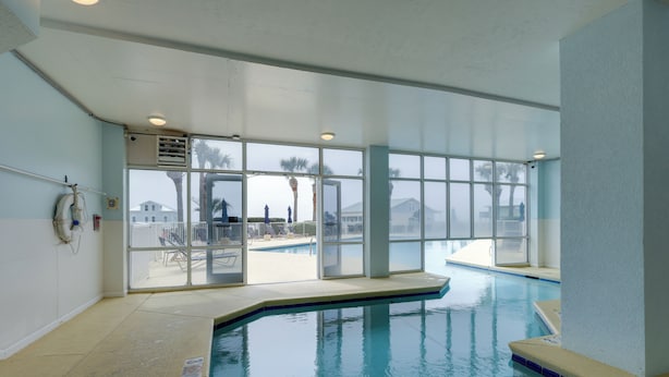 Ein Hallenbad mit großen Glaswänden, einem Rettungsschwimmerstand und Blick auf den Außenbereich des Schwimmbads.
