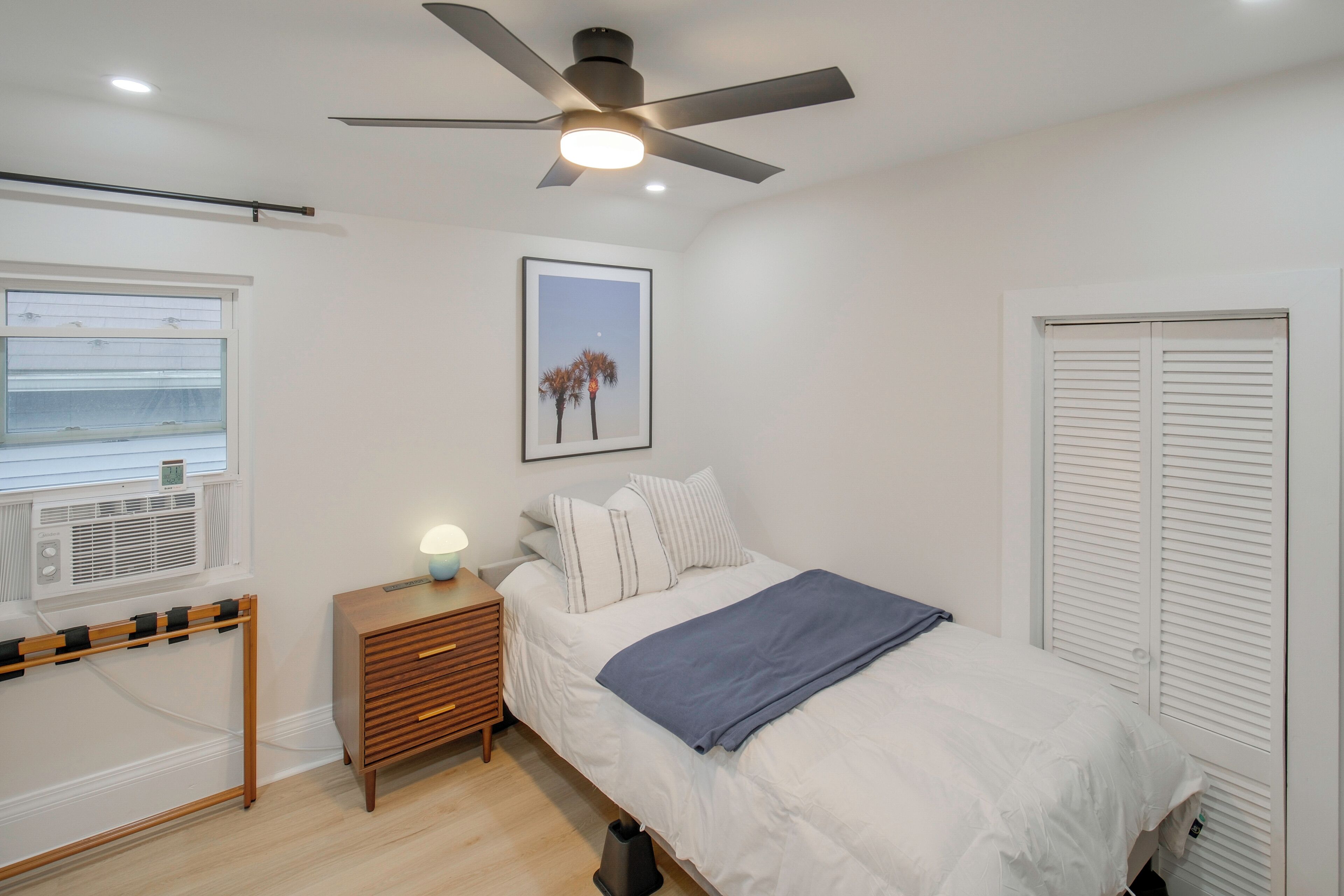 A bedroom with a bed, bedside table, ceiling fan, window, air conditioning unit, and a framed picture on the wall.