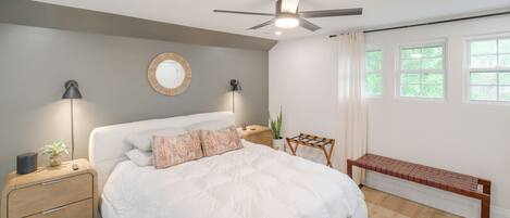 A bedroom with a bed, bedside tables, a ceiling fan, a round mirror, and a window with curtains.