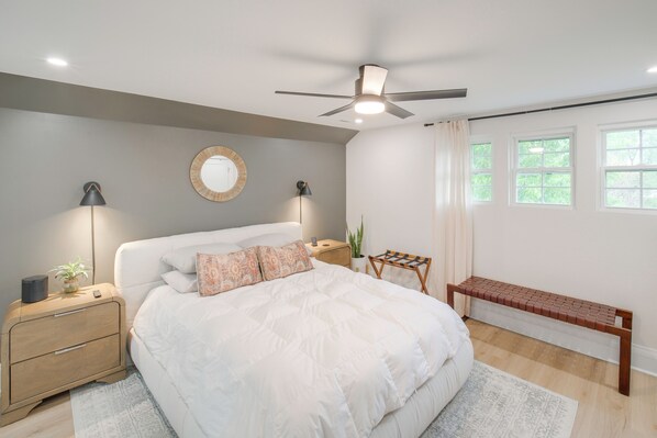 Une chambre à coucher comprenant un lit, des tables de chevet, un ventilateur de plafond, un miroir rond et une fenêtre avec des rideaux.
