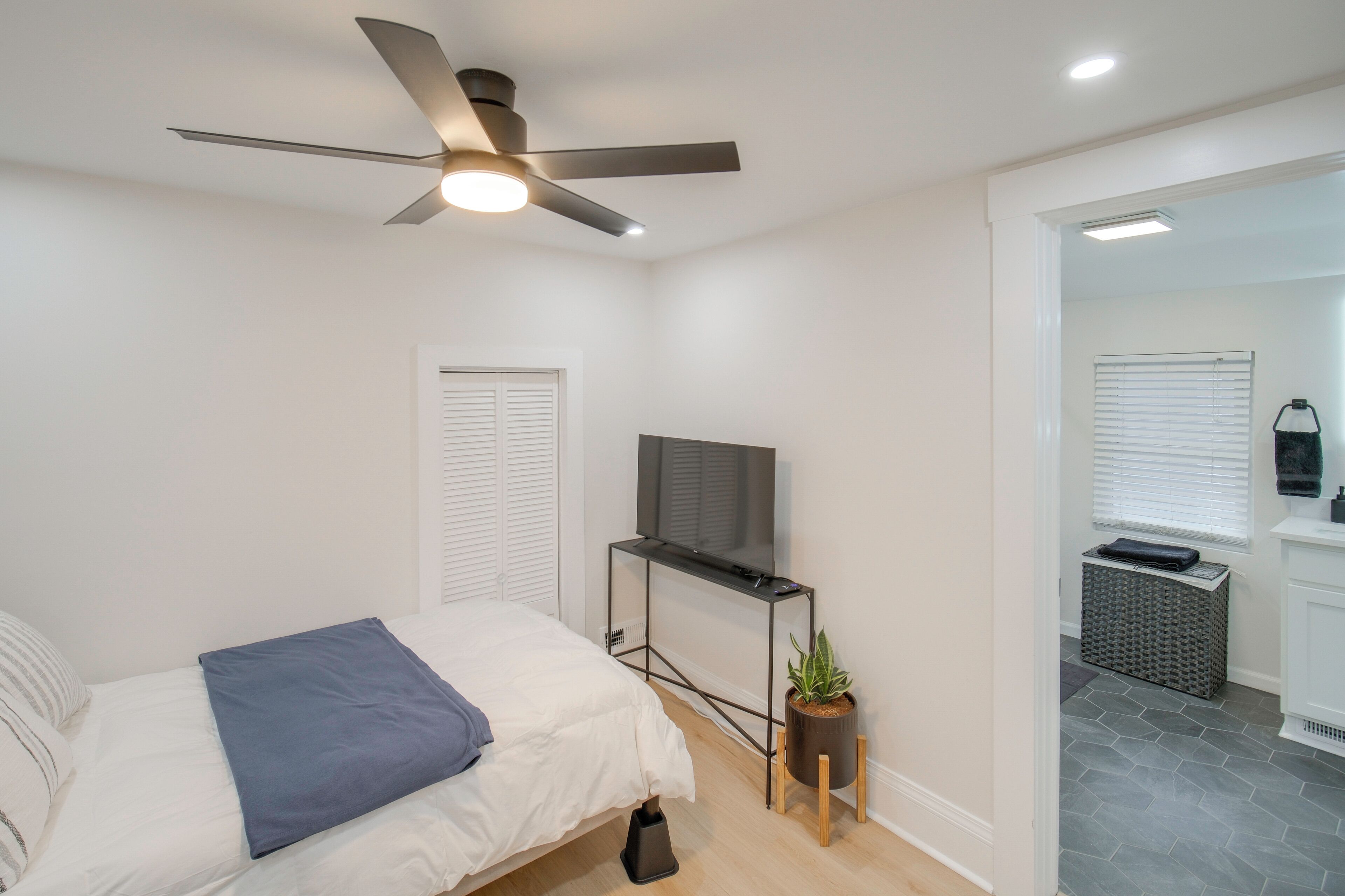 A bedroom with a bed, a ceiling fan, a TV on a stand, a small plant, and a bathroom visible through an open door.