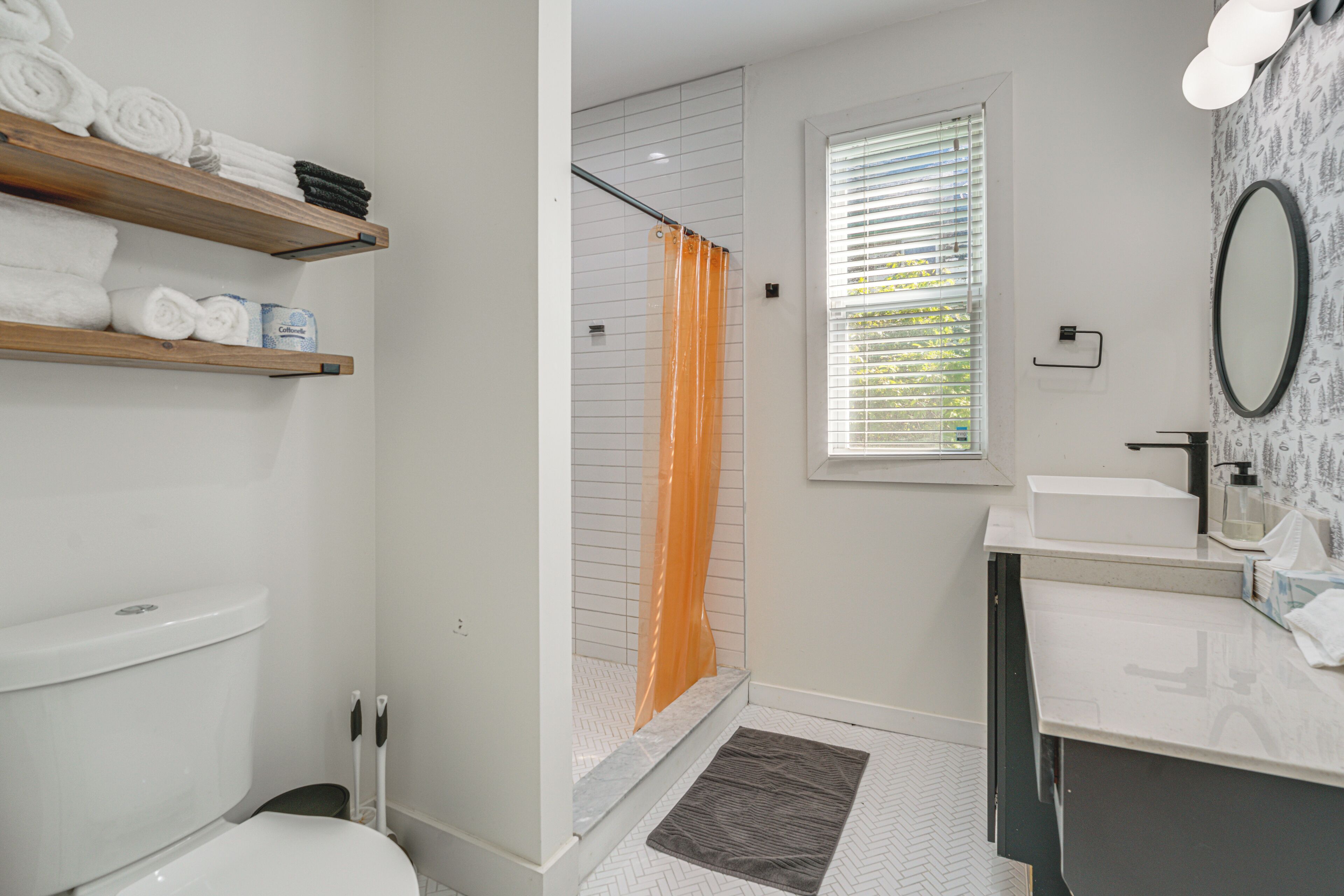A bathroom with a toilet, a shower with an orange curtain, a sink with a mirror, and shelves with towels.