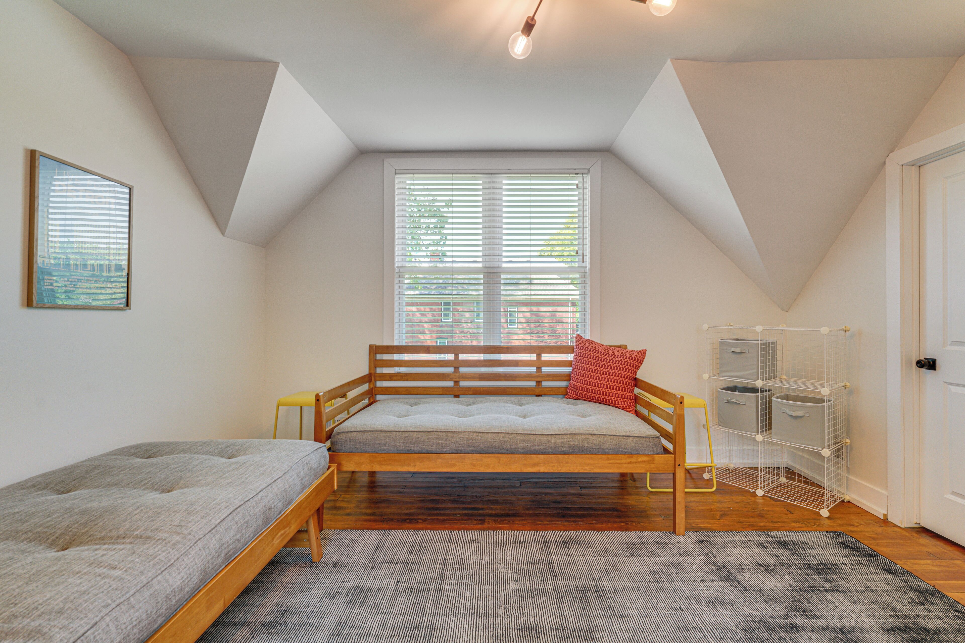 Une chambre située dans un espace de type « loft », avec un plafond en pente, un lit doté d’une tête de lit, une fenêtre avec des stores, une porte et un meuble de rangement.