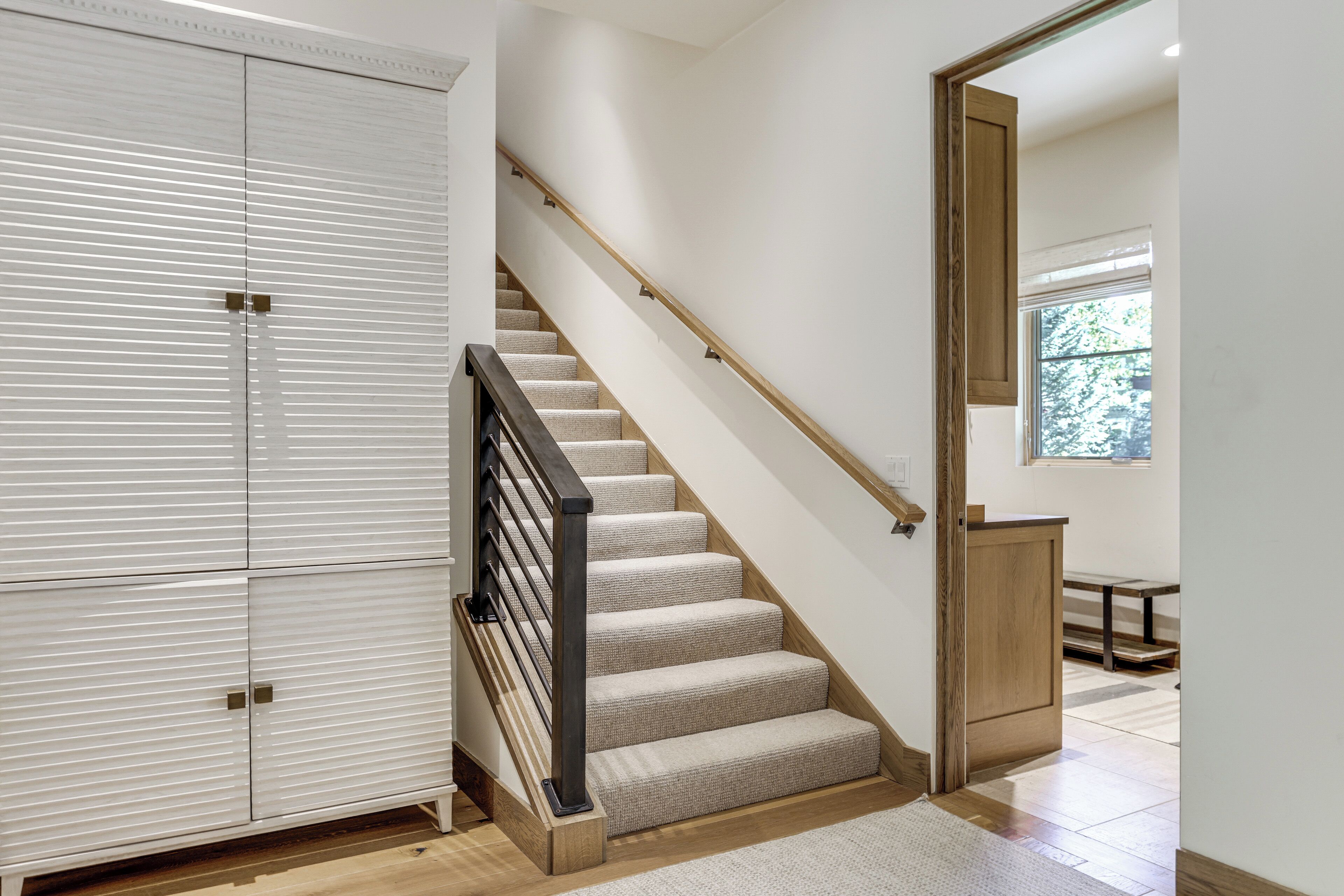 A staircase with a wooden handrail, beige carpeted steps, and a white wall with a window.
