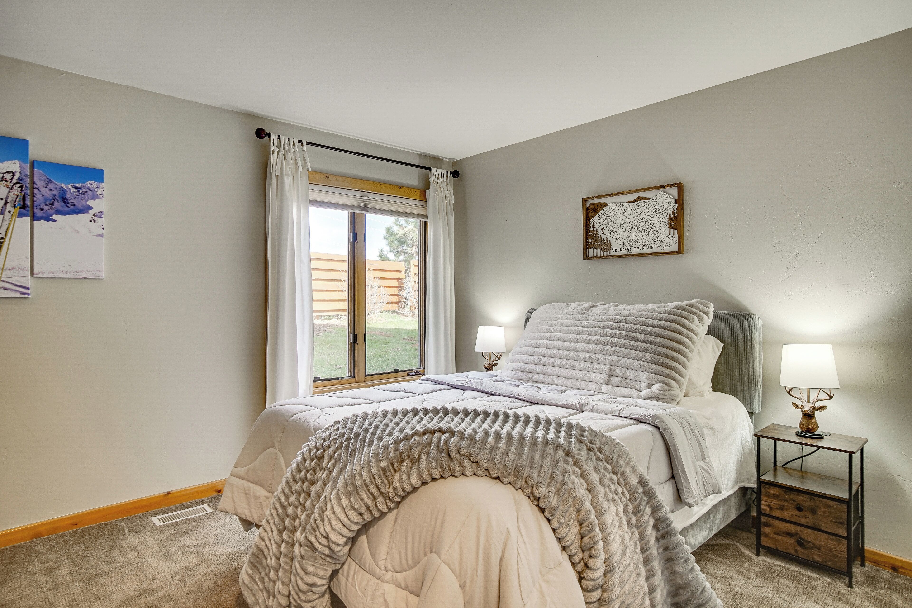 Quarto com cama, duas mesas de cabeceira com abajures, janela com cortinas e um quadro na parede.
