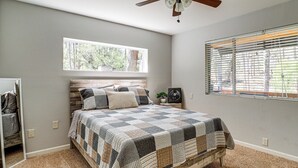 A bedroom with a wooden bed, a large window with blinds, a ceiling fan, and a nightstand with a lamp. - Deck, Trails & Forest Views! Ponderosa Park Cabin (Prescott)
