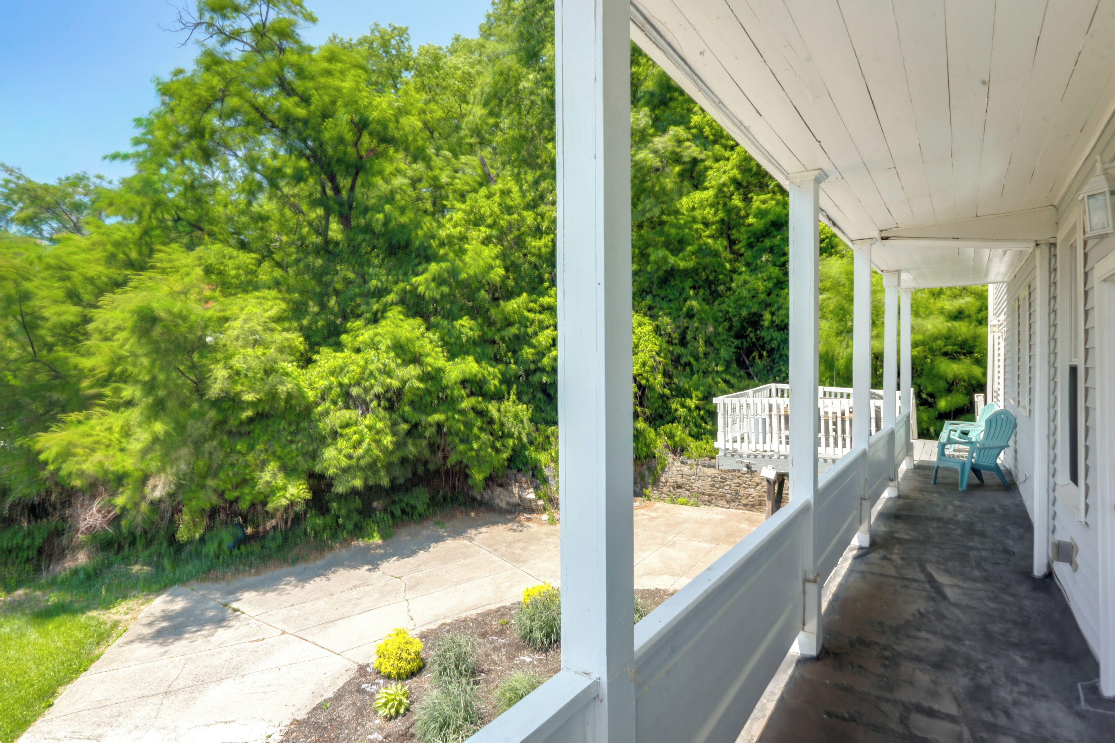 A covered porch with white railings, a stone patio, and a view of a lush green landscape.