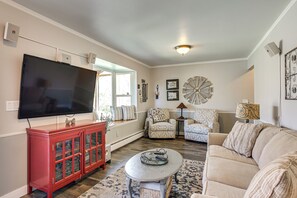 Una sala de estar con televisor de pantalla plana, un mueble de almacenamiento rojo, un sillón estampado, un sofá color crema y un reloj de pared decorativo.