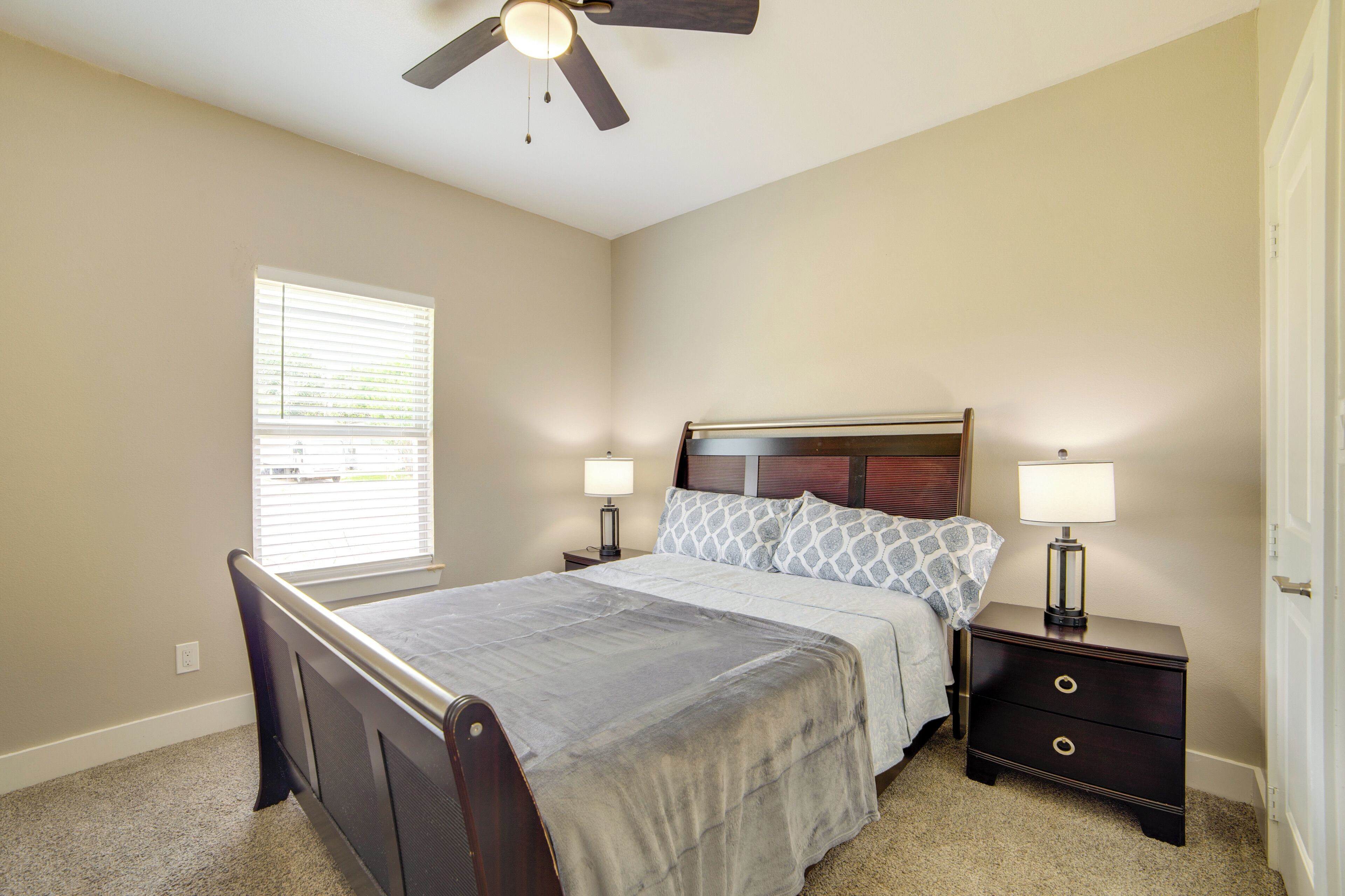 A bedroom with a bed, two bedside tables with lamps, a ceiling fan, and a window with blinds.
