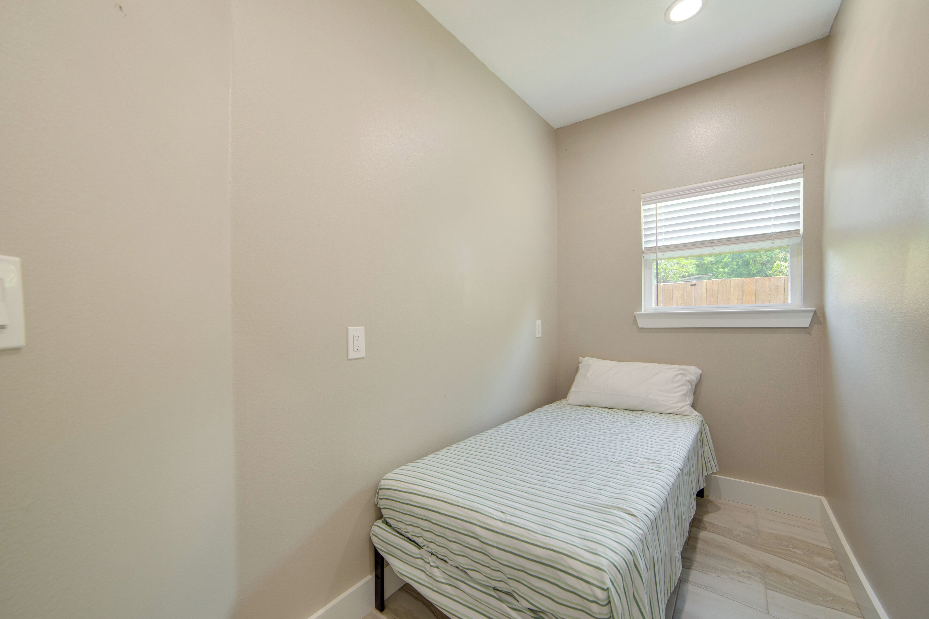 Une petite chambre avec un lit simple, une fenêtre, un luminaire au plafond et un plancher en bois.