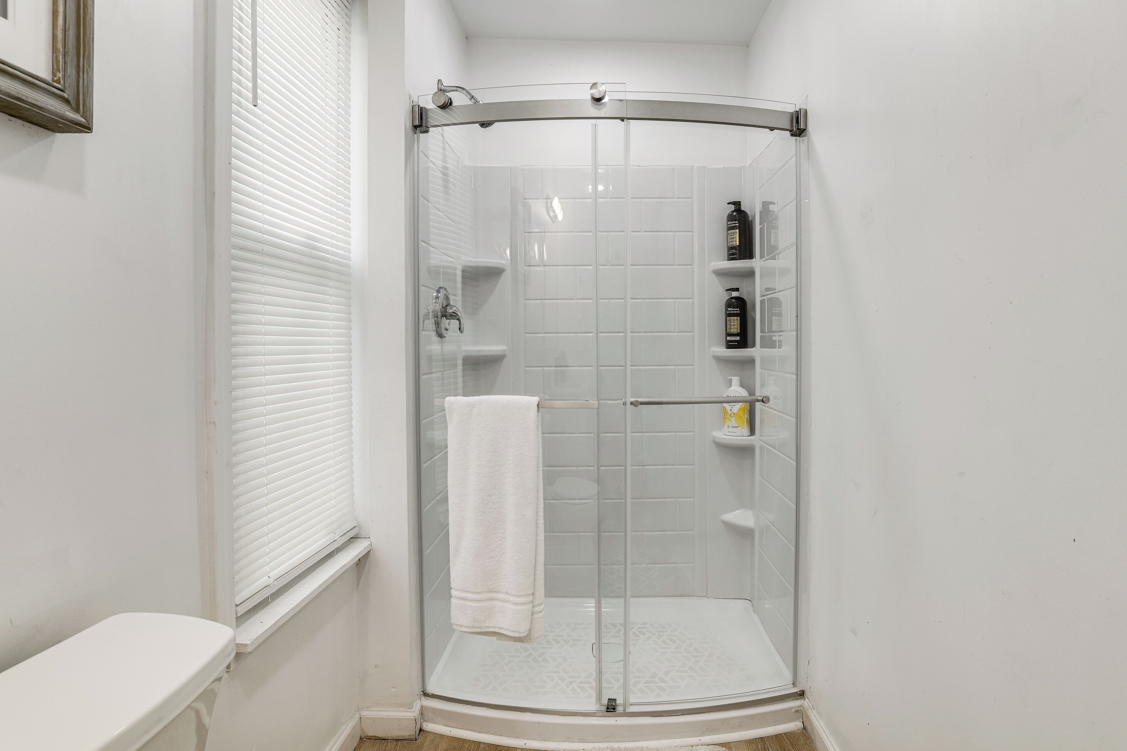 A corner shower with a glass door, white tiled walls, a white towel hanging on the door, and a toilet to the left.