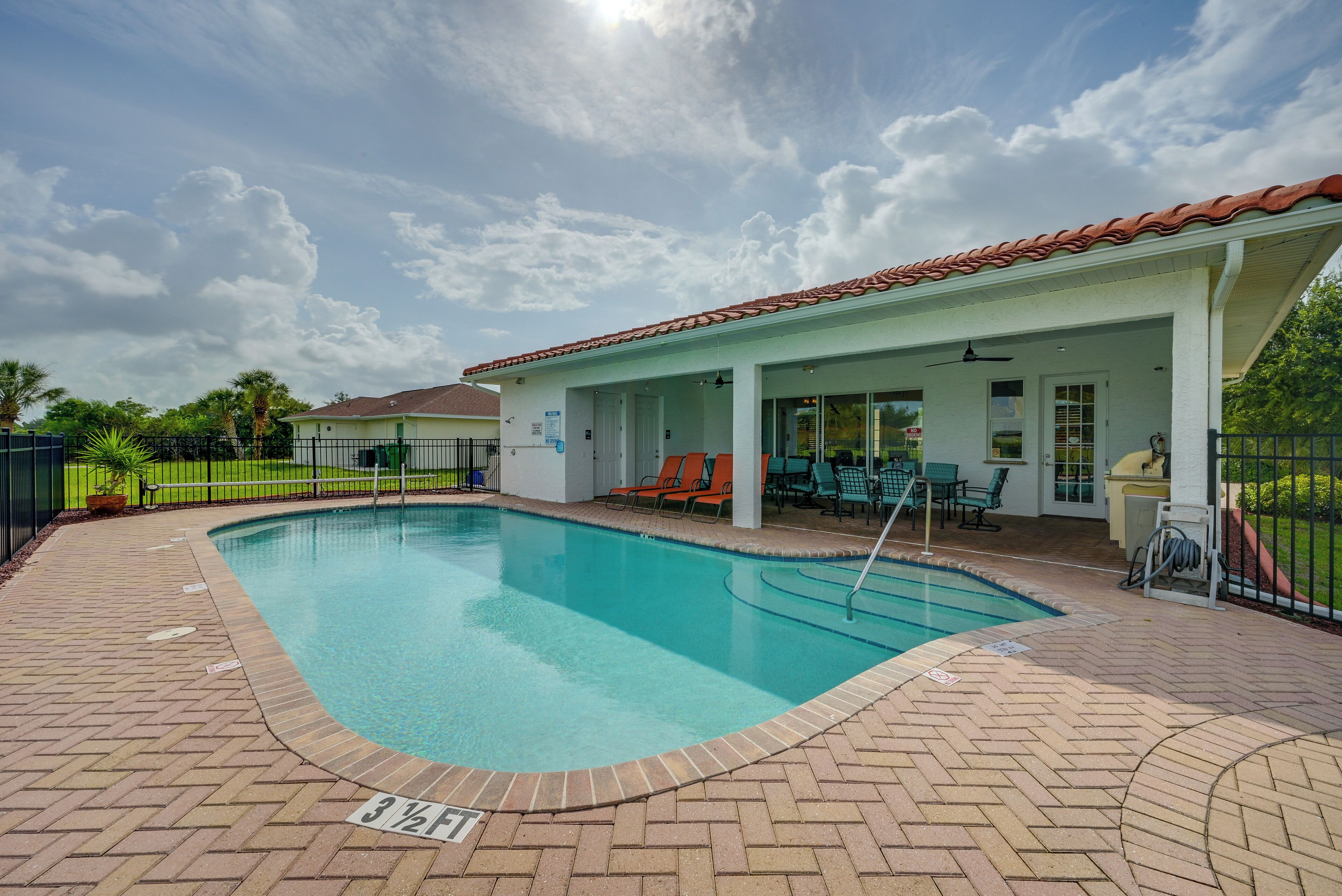 En swimmingpool med brobelagt terrasse, et overdækket opholdsområde med stole, og en klar himmel.