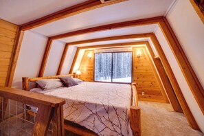 A bedroom with a sloped ceiling, a bed with a patterned bedspread, a window with a view of trees, and wooden walls. - 4-cabin Haven Near Kenai River: Catch & Freeze! (Sterling)