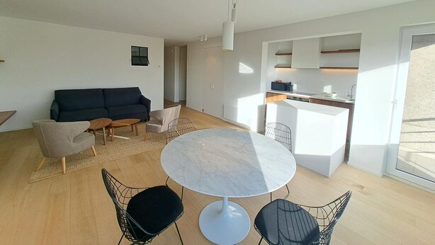A modern living space with a round marble table, a dark sofa, and a kitchen area with white cabinets. - Sunny Apartment Near the Beach (Koksijde)