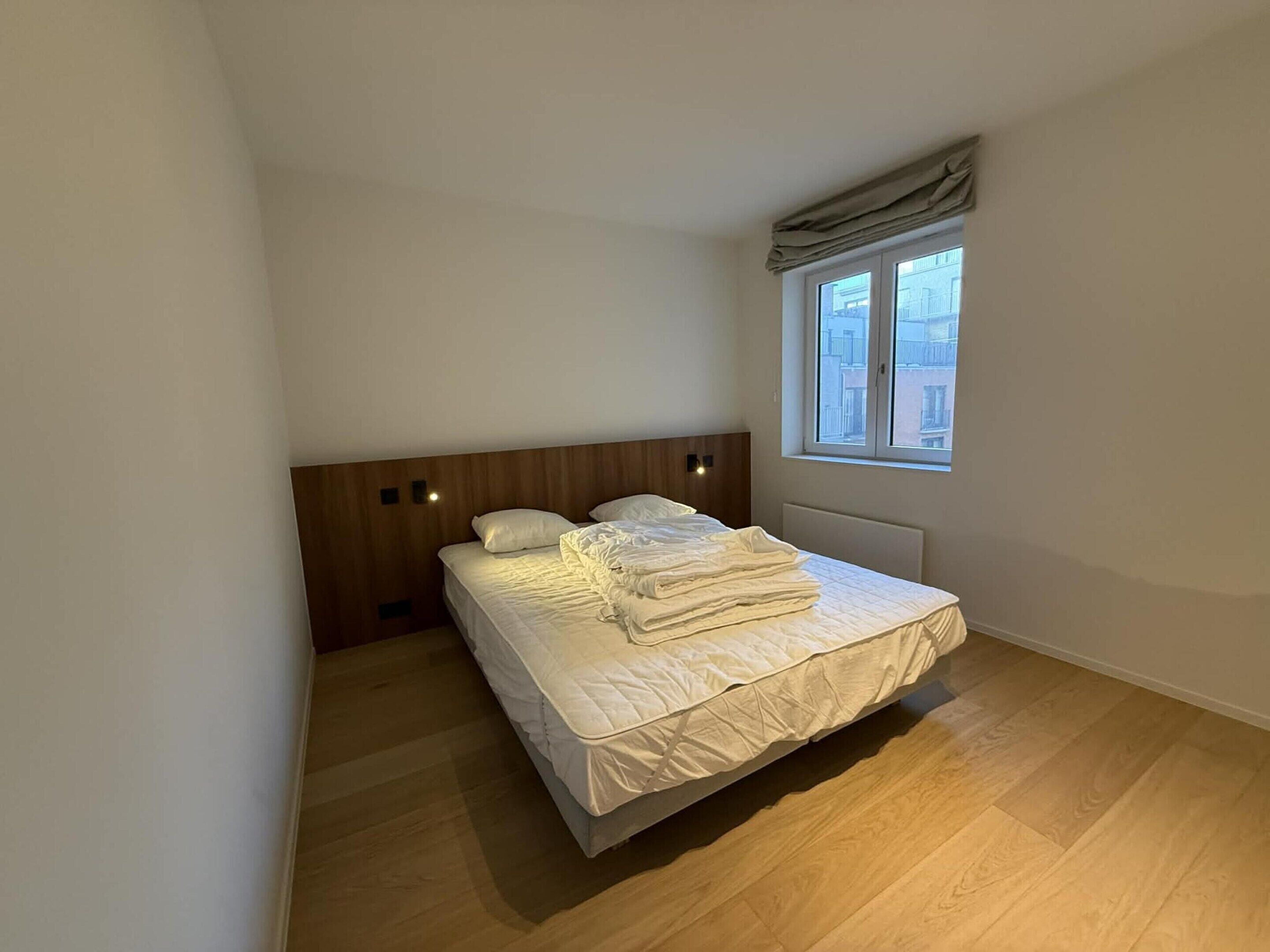 Ein kleines, ordentlich eingerichtetes Schlafzimmer mit einem Holzkopfende, einem Bett mit weißer Bettwäsche, einem Fenster mit Blick auf Gebäude und einem Heizkörper unter dem Fenster.