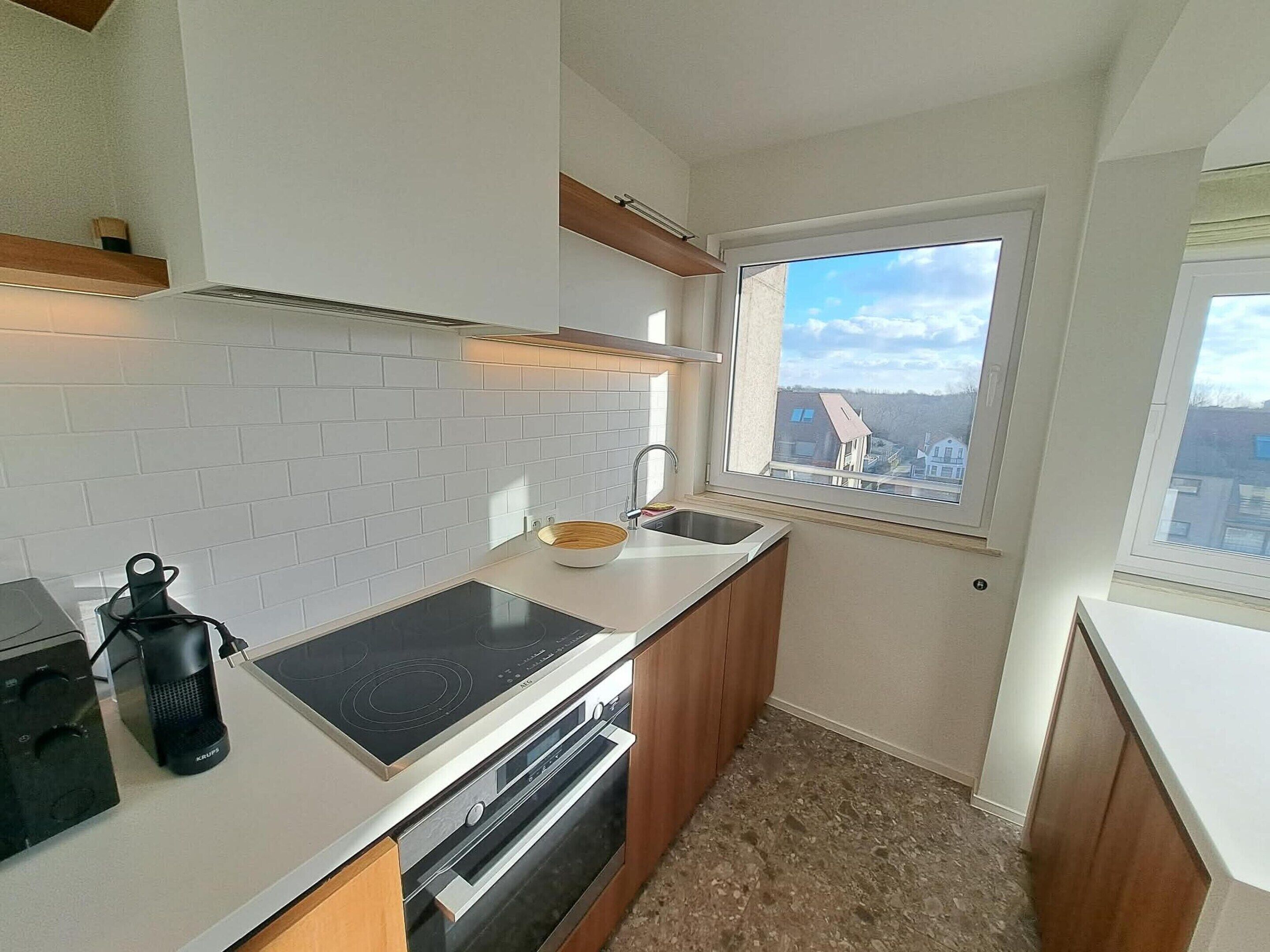 A compact kitchen with white tiles, a built-in stove, a sink, and a window with a view of buildings.