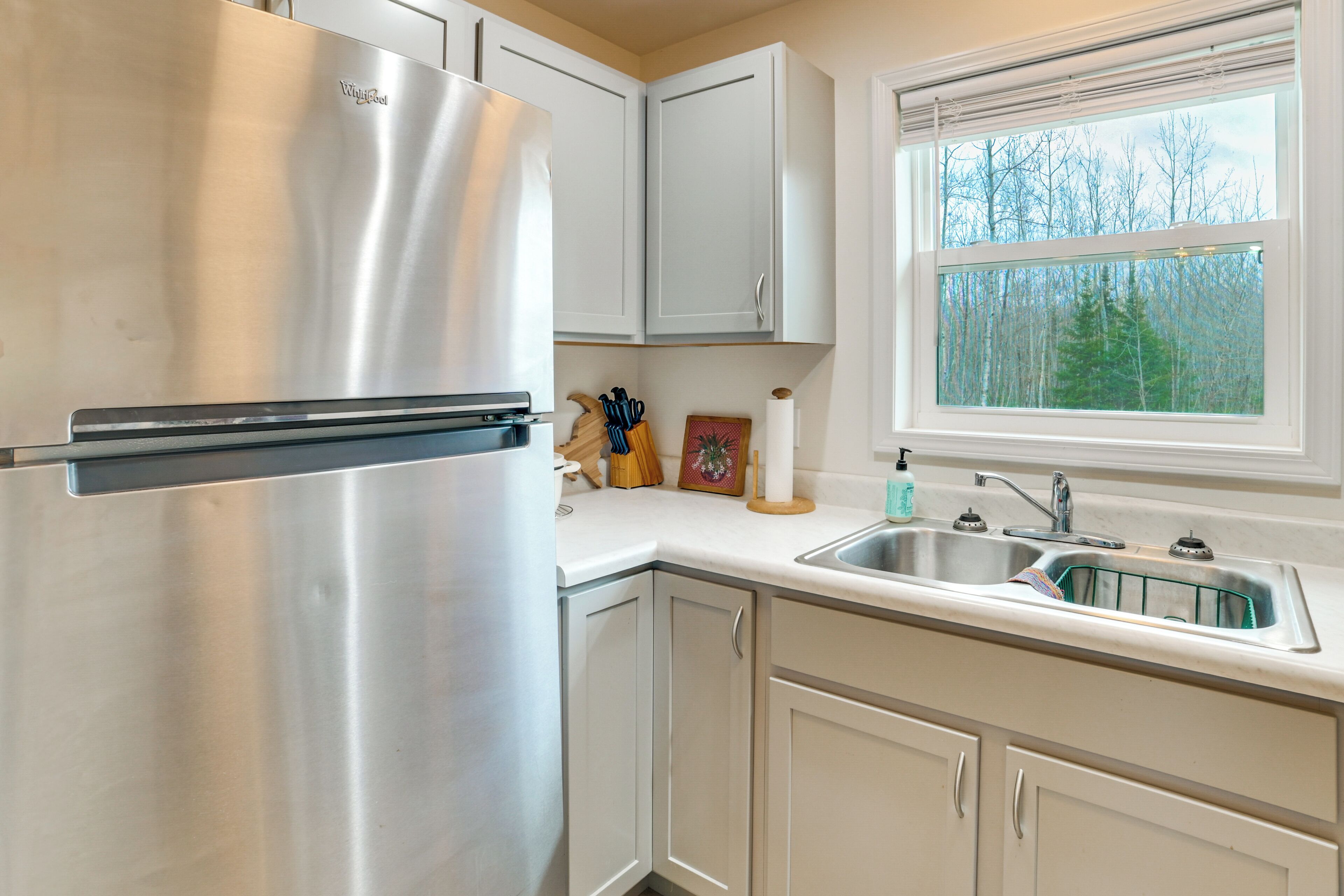 Una cocina con un refrigerador de acero inoxidable, armarios blancos, un ventanal con vista a árboles y un lavamanos con grifo.