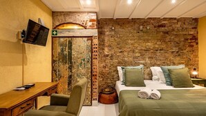 Ett rum med en rustik tegelvägg, en säng med grönt sänglinne, ett skrivbord i trä och en tv som är monterad på väggen.