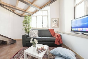 A living room with a black leather sofa, a white coffee table with books and flowers, a TV on the wall, a staircase, and a large window with curtains. - Captivating Loft in Prime Location Near Colosseum (Rome)