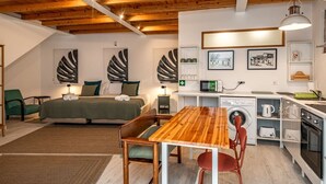 Ein gemütliches Schlafzimmer mit Bett, Nachttischen und einem grünen Sessel. In der Mitte steht ein Holztisch mit Stühlen. Eine Küchenzeile mit Mikrowelle, Wasserkocher und Backofen befindet sich rechts.