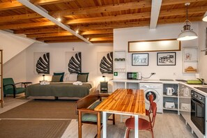 A compact bedroom with a bed, bedside tables, and a green chair. A wooden dining table is in the center with chairs. A kitchenette with a microwave, kettle, and oven is on the right. - Secret Riverside Studio in Porto Brandão (Caparica)