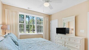 Quarto com cama, penteadeira, televisão e janela com cortinas.